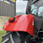 Close-up of a red tractor hood with a large black tire, parked beside a gray building with signboards and a stone wall to the left.
