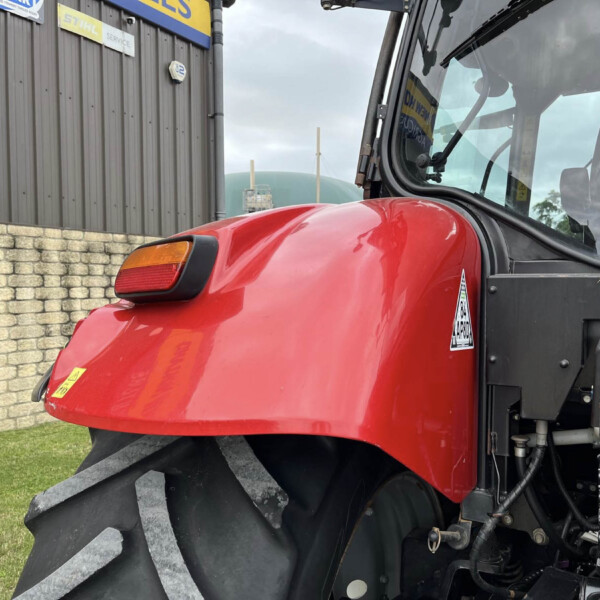 Close-up of a red tractor hood with a large black tire, parked beside a gray building with signboards and a stone wall to the left.