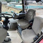Cab interior of a construction vehicle showing steering wheel, pedals, and gray fabric seat; a worksite with blue-covered ground outside the window.