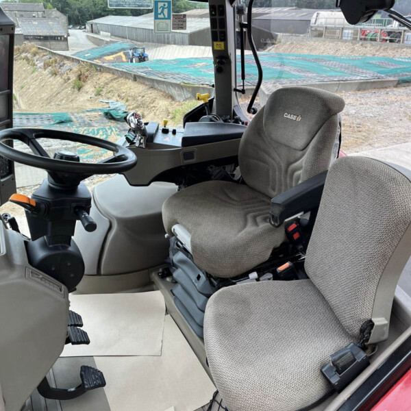 Cab interior of a construction vehicle showing steering wheel, pedals, and gray fabric seat; a worksite with blue-covered ground outside the window.