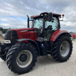 Close-up of a red tractor with oversized front and rear tires in a gravel lot under a cloudy sky.