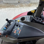 Inside a tractor cab showing a control panel with many colored buttons and two yellow joystick levers, red exterior hood visible outside the window.