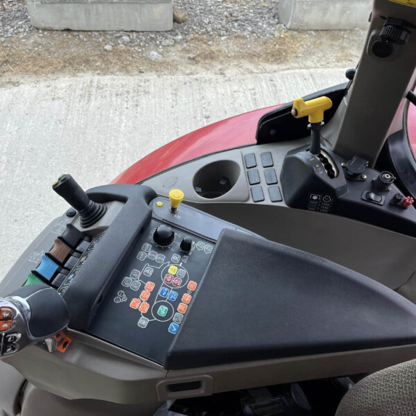 Inside a tractor cab showing a control panel with many colored buttons and two yellow joystick levers, red exterior hood visible outside the window.