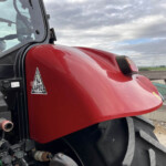 Close-up of a red tractor hood and cabin window with hydraulic hoses and controls visible at the bottom left, on a farm.