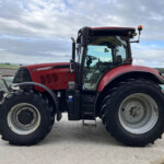 Red farm tractor with large tires, side view, parked on a concrete surface under a cloudy sky, cab visible.