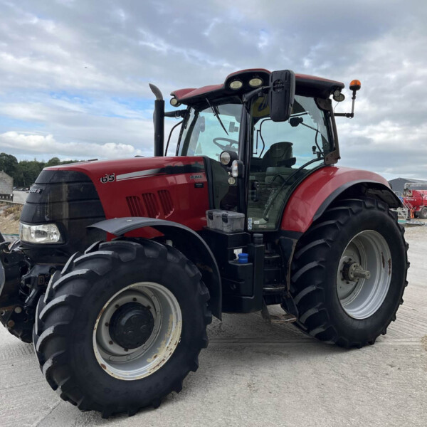 Close-up of a red tractor with oversized front and rear tires in a gravel lot under a cloudy sky.