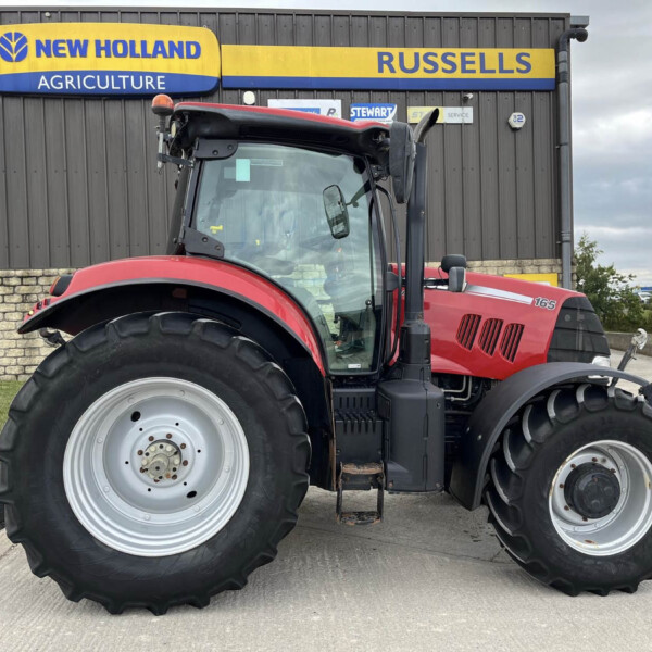 Red agricultural tractor with oversized front and rear tires parked in front of a gray industrial building with a New Holland Agriculture sign