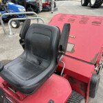 Red ride-on lawn tractor with black seating area, parked in a lot with blue tractors in the background