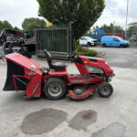 Red ride-on lawn mower in the foreground at an industrial yard with various equipment and a metal gate behind it.