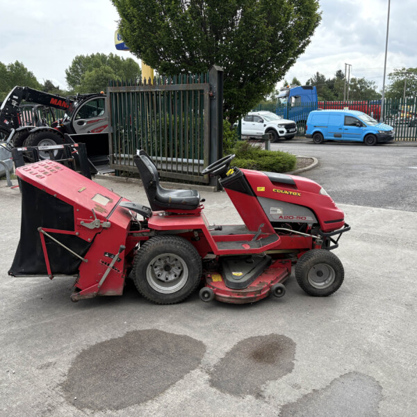 Red ride-on lawn mower in the foreground at an industrial yard with various equipment and a metal gate behind it.