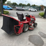 Red ride-on lawn mower with a large black attachment in a parking lot near a green fence.