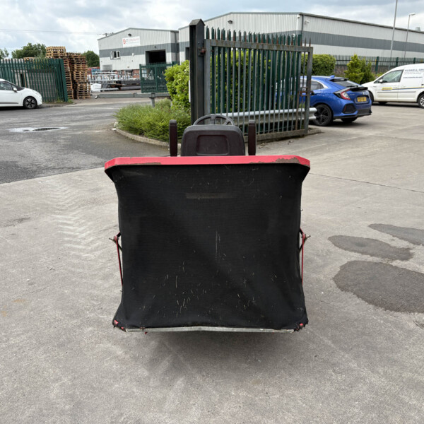 View from behind a large black and red waste container in a parking lot outside an industrial warehouse, with pallets, fencing, and parked cars visible in the background.
