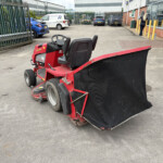 Red riding street-sweeper with a large black plow in a parking lot near a building and fence.