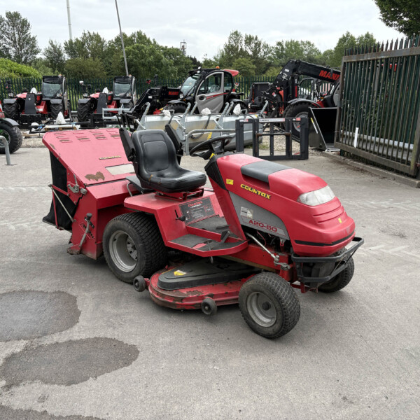 Red ride-on lawn mower in the foreground with row of tractors and equipment behind a fence.