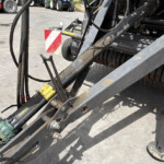 Close-up of a dusty industrial machine frame with hoses, metal beams, and a red‑and‑white safety reflector; warning stickers visible in the scene.