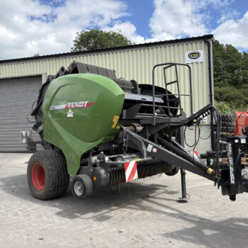 Green Fendt combine harvester on a trailer parked in front of a beige corrugated metal warehouse, with red farm machinery nearby under a blue sky.
