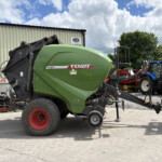 Green Fendt 4160 V round baler connected to a tractor in an outdoor equipment yard near a corrugated metal shed under a cloudy sky.