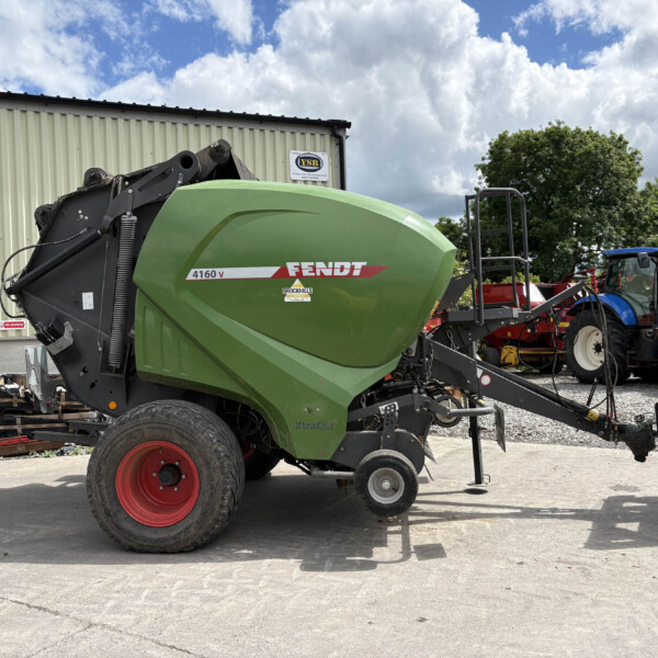 Green Fendt 4160 V round baler connected to a tractor in an outdoor equipment yard near a corrugated metal shed under a cloudy sky.