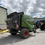 Green Fendt agricultural combine parked outdoors beside a beige corrugated metal building under a cloudy sky, in a yard with other machinery nearby.