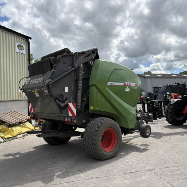 Green Fendt agricultural combine parked outdoors beside a beige corrugated metal building under a cloudy sky, in a yard with other machinery nearby.