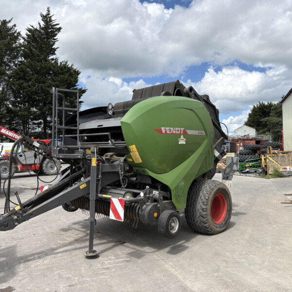 Green Fendt round baler in a farm equipment yard with other machinery and stacks of tires nearby, under a partly cloudy sky.