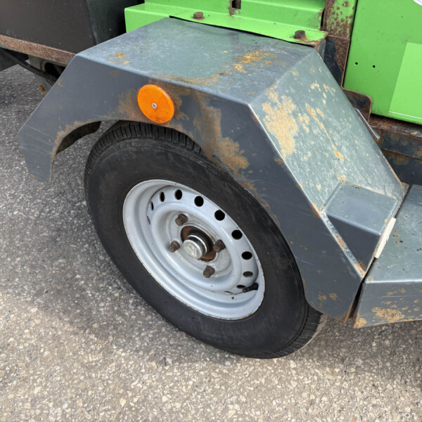 Close-up of a rusted trailer fender over a black tire with an orange side marker light, green equipment in the background.