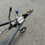 Close-up of a trailer hitch assembly on a metal frame, featuring a blue cap on the jack and a black accordion dust boot, set on an asphalt surface.