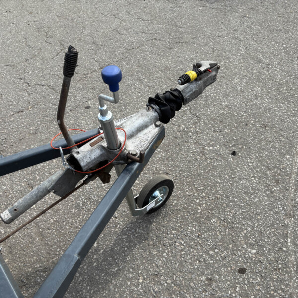 Close-up of a trailer hitch assembly on a metal frame, featuring a blue cap on the jack and a black accordion dust boot, set on an asphalt surface.