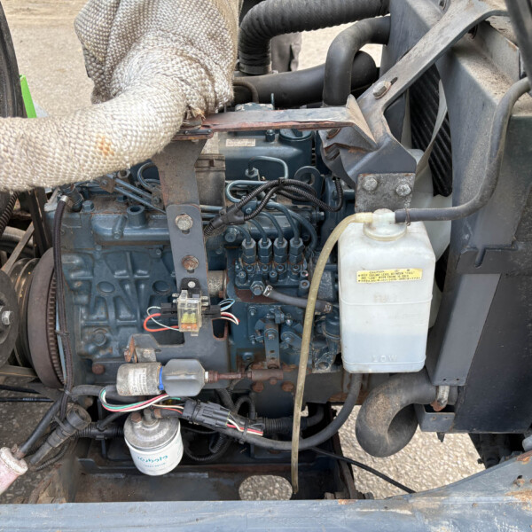 Tractor engine bay with blue engine block, wiring, hoses, and a gloved hand resting on a metal bracket amidst dusty components