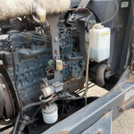 Close-up of a blue industrial engine in a rusted metal frame, with belts, hoses, and a white coolant reservoir visible inside the machine.