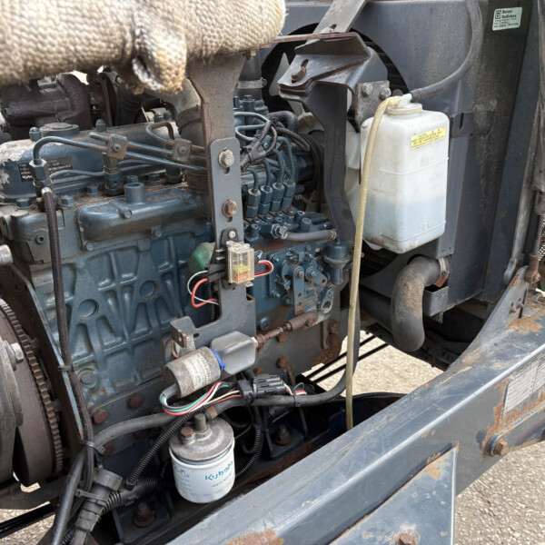 Close-up of a blue industrial engine in a rusted metal frame, with belts, hoses, and a white coolant reservoir visible inside the machine.