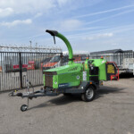 Green Arborist 200 wood chipper on a trailer in an industrial yard behind a tall metal fence with barbed wire under a blue sky with clouds.