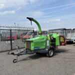 Green Arborist 200 wood chipper on a trailer in a fenced industrial yard under a blue sky and clouds, barbed wire fence in foreground.