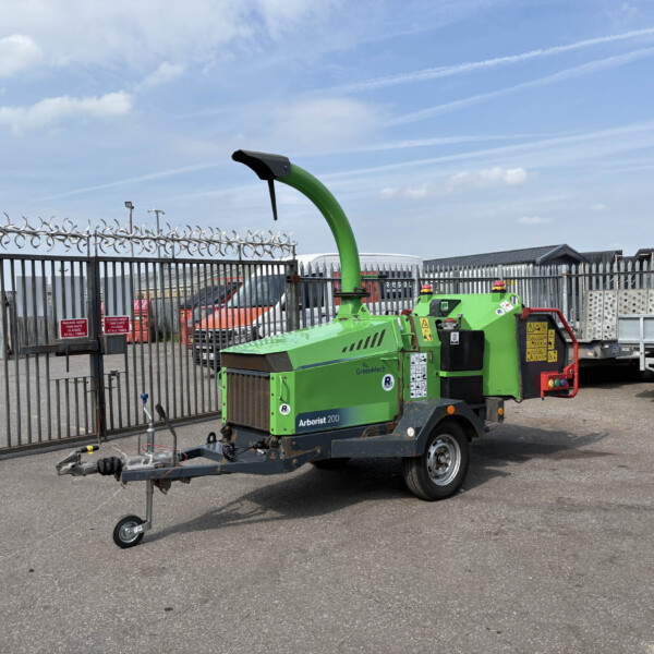 Green Arborist 200 wood chipper on a trailer in a fenced industrial yard under a blue sky and clouds, barbed wire fence in foreground.