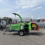 Green arborist wood chipper on a trailer in an industrial yard, with metal fencing and parked vans under a blue sky.