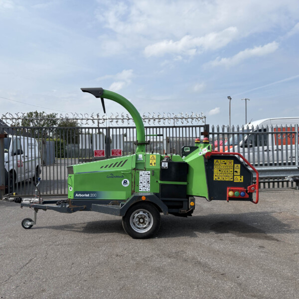 Green arborist wood chipper on a trailer in an industrial yard, with metal fencing and parked vans under a blue sky.
