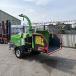 Green agricultural machine in an outdoor yard with a tall metal fence and barbed-wire scene edge-to-edge on a sunny day