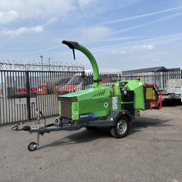Green Arborist 200 wood chipper on a trailer in an industrial yard behind a tall metal fence with barbed wire under a blue sky with clouds.