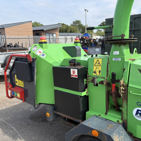 Green heavy-duty machine in an outdoor storage yard, with a metal fence, portable buildings, and pallets in the background.