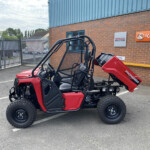 Red off-road utility vehicle with roll cage parked in a parking lot beside a brick building with Kubota and Honda signage in view.
