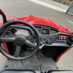 Driver’s view from a red utility vehicle: steering wheel, dashboard screen, and textured floor mats with pedals visible; outside is a paved street and gate.