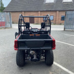 Rear view of a red Honda side-by-side utility vehicle with a cargo bed and roll cage in a parking lot, brick building in the background.