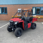 Red Honda Pioneer utility vehicle parked in a marked parking space in front of a brick building with blue-framed windows.