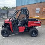Red Pioneer side-by-side utility vehicle parked in a lot outside a brick building with Honda and Kubota signs nearby, showing roll cage and cargo bed
