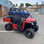 Red off-road utility vehicle with a black roll cage and mesh seating in a parking lot by a brick building and warehouse.