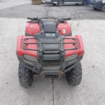 Front view of a red Honda ATV parked on a concrete lot with muddy tires and black handlebars, Trucks visible in the background.