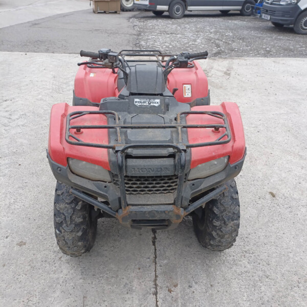 Front view of a red Honda ATV parked on a concrete lot with muddy tires and black handlebars, Trucks visible in the background.