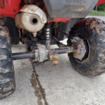 Rear underside of a muddy red quad bike, showing exhaust, suspension, and chunky off-road tires.