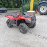 Red ATV parked on a concrete surface with a large blue tractor visible in the background and greenery beyond.