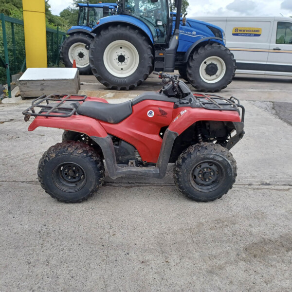 Red Honda ATV in foreground with muddy tires on a concrete lot; blue tractors and a New Holland Agriculture van in the background near a green fence.
